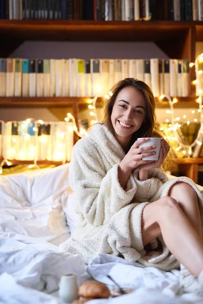 Girl in white warm bathrobe sitting on the bed with glowing christmas garland lights on background and having breakfast — Stock Photo, Image