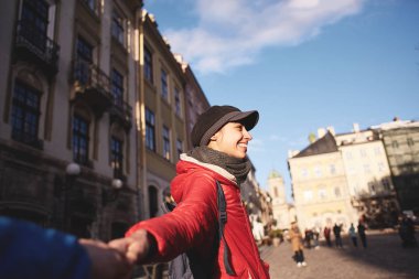 ilk kişi görünümü kırmızı ceket, gri eşarp ve kap içinde bir genç gülümseyen kadın. Yürüyen kadın açık havada güneşli Güz veya bahar günü ve ilan, sokakta birisi elle beni izleyin kavramı
