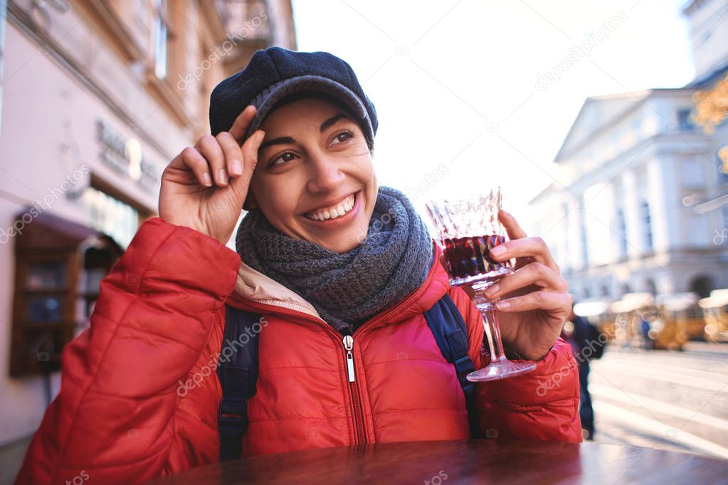 Image of smiling young woman standing outdoors in city street cafe holding glass drinking wine