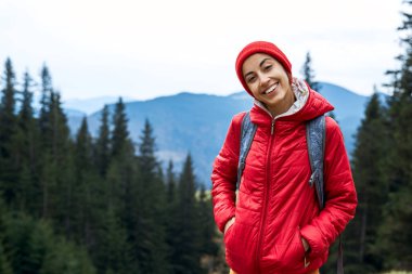 kırmızı ceket ve kap, orman ve dağlar arka plan üzerinde duran giyen kadın uzun yürüyüşe çıkan kimse ile sırt çantası,