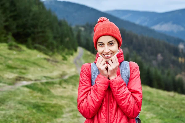 Mujer excursionista con mochila, con chaqueta roja y pantalones anaranjados, de pie sobre el fondo de las montañas —  Fotos de Stock