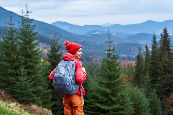 Mulher caminhante com mochila, vestindo jaqueta vermelha e calças laranja, de pé sobre o fundo das montanhas — Fotografia de Stock