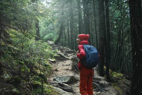 Escursionista donna con zaino, indossando giacca rossa e pantaloni arancioni, passeggiando nel bosco in montagna — Foto Stock