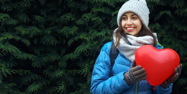 happy smiling young woman in warm jacket, knitted cap and scarf is standing outdoors with a heart shaped red balloon