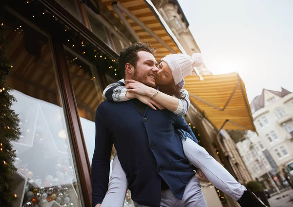 Feliz pareja enamorada está de pie al aire libre cerca de los escaparates decorados, abrazándose y besándose —  Fotos de Stock