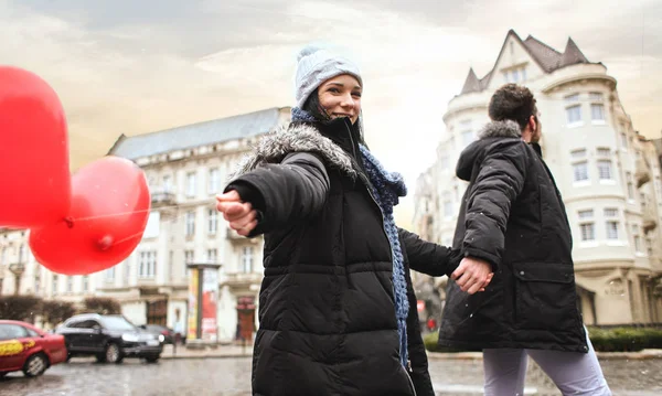 Feliz riendo pareja en caliente invierno ropa es caminar al aire libre con un corazón en forma de globos rojos —  Fotos de Stock