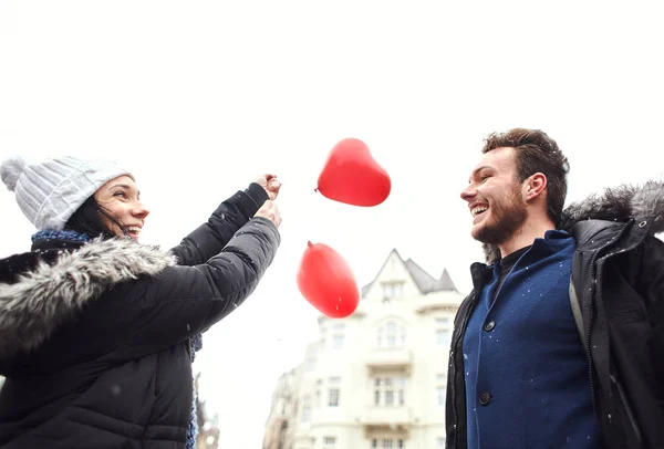 Feliz riendo pareja en caliente invierno ropa es caminar al aire libre con un corazón en forma de globos rojos —  Fotos de Stock