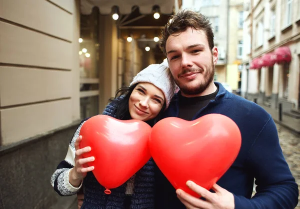 Feliz riendo pareja en caliente invierno ropa es caminar al aire libre con un corazón en forma de globos rojos —  Fotos de Stock