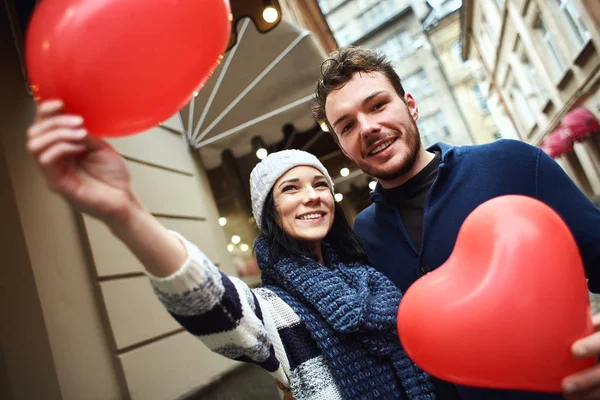 Счастливая смеющаяся пара в уютных зимних свитерах стоит на улице с красными шарами в форме сердца в руках — стоковое фото