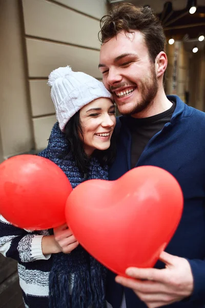 Lyckligt skrattande par i mysig vinter tröjor står utomhus med en hjärtformade röda ballonger i händerna — Stockfoto