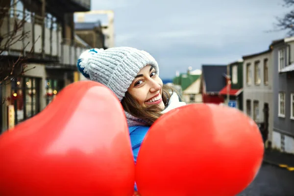 happy young woman in warm winter clothes, knitted cap and scarf is standing outdoors with a heart shaped red balloons