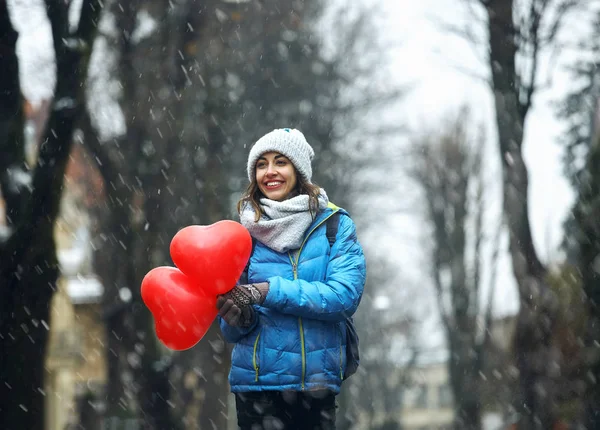Mujer joven feliz en ropa de invierno caliente, gorra de punto y bufanda está de pie al aire libre con un corazón en forma de globos rojos —  Fotos de Stock