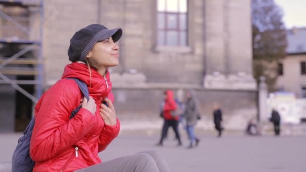 Mujer joven con chaqueta roja y gorra gris sentada en la calle — Vídeos de Stock