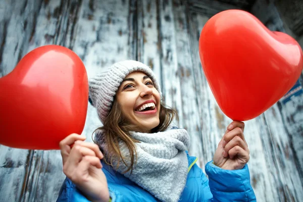 happy young woman in warm winter clothes, knitted cap and scarf is standing outdoors with a heart shaped red balloons