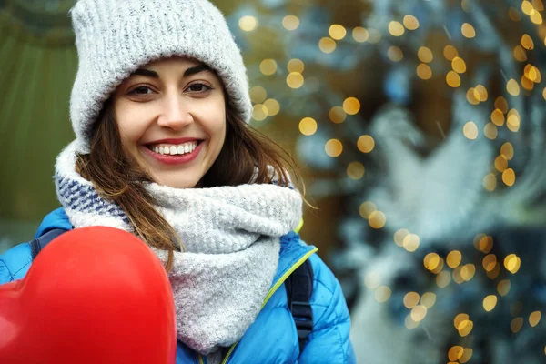 Щаслива молода жінка в теплому зимовому одязі, в'язана шапка і шарф стоїть на відкритому повітрі з червоними кульками у формі серця — стокове фото