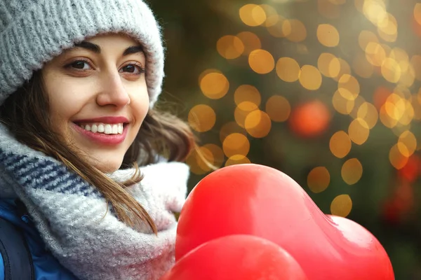 Felice giovane donna in abiti invernali caldi, cappello a maglia e sciarpa è in piedi all'aperto con un palloncini rossi a forma di cuore — Foto Stock