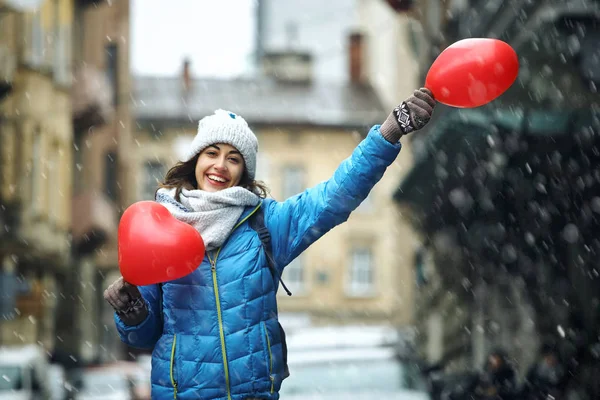 Happy young woman in warm winter clothes, knitted cap and scarf is standing outdoors with a heart shaped red balloons — Zdjęcie stockowe