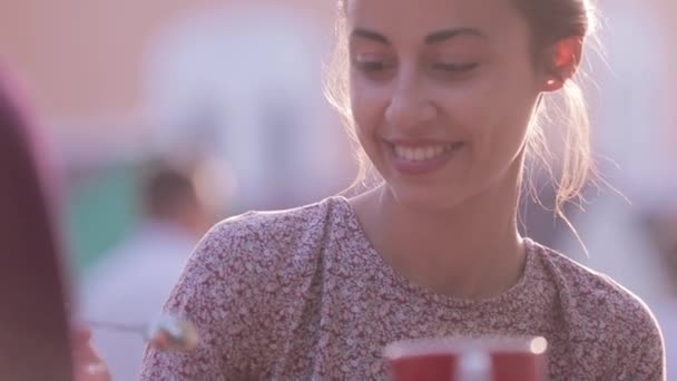 Hermosa mujer está sentada al aire libre en la mesa con tazas de café, hablando con alguien y mirando hacia atrás — Vídeo de stock