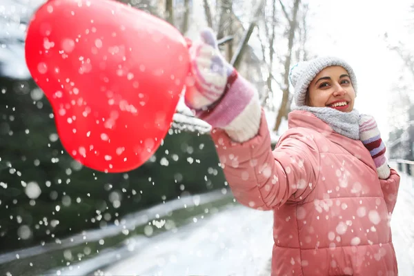 Wizerunek szczęśliwy radosny kobiety uśmiechający się szczęśliwie chodzenie na zewnątrz w mroźny zimowy śnieżny dzień z sercem w kształcie czerwony balonik — Zdjęcie stockowe
