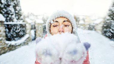 sıcak kış giysileri içinde sevimli mutlu neşeli gülümseyen kadın hem avuç içi bir kar tutan ve kameraya kar üfleme