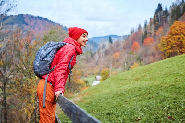 Женщина турист с рюкзаком, одет в красную куртку и оранжевые брюки, стоя на фоне гор — стоковое фото