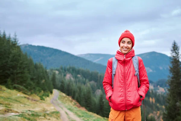 Mulher caminhante com mochila, vestindo jaqueta vermelha e calças laranja, de pé sobre o fundo das montanhas — Fotografia de Stock