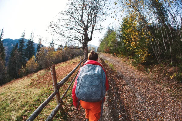 Woman hiker with backpack, wearing in red jacket and orange pants, going up on the mountain road in mountains — Stock Photo, Image