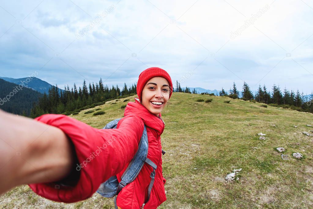 selfie image of a young smiling woman hiker with small backpack standing on a mountain slope.