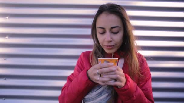 Young woman in red jacket with blue jeans Waist bag drinking coffee outdoors on a metal wall background — Stock Video