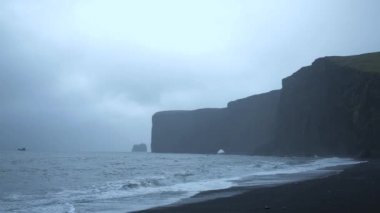 Izlanda 'da Reynisfjara siyah kum Plajı yakınında kayalıklar, kayalar, okyanus ve karanlık yağmur ve fırtına bulutları ile siyah kum plaj Panoraması