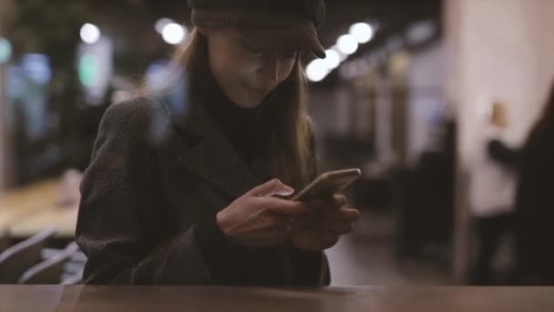 Portret młodej pięknej modnej kobiety brunetka siedzi w kawiarni w pobliżu okna wieczorem i za pomocą telefonu. Modelka ubrana w stylowy szary płaszcz, kapelusz i czarny Golf. Sezonowa Moda kobieca — Wideo stockowe