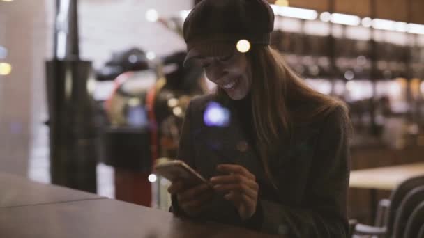 Portret van jonge mooie modieuze brunette vrouw zittend in coffeehouse buurt van het venster 's avonds en het gebruik van telefoon. Model dragen stijlvolle grijze jas, hoed en zwarte golf. Seizoensgebonden vrouwelijke mode — Stockvideo