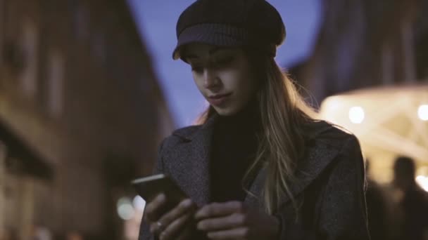 Портрет молодої красивої модної брюнетки, що стоїть на вулиці ввечері і використовує телефон. Модель в стильному сірому пальто, капелюсі та чорному гольфі. HD стокове зображення — стокове відео