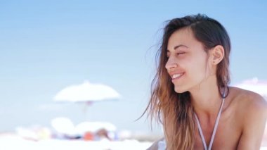 güzel bronzlaşmış gülümseyen kadın beyaz kum sahilde yatıyor, rahatlatıcı ve küçük kulaklıklar ile telefondan müzik dinlemek. Bikini ve beyaz kap üzerinde ince seksi kadın enjoing ve güneş