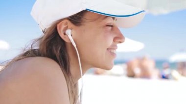 güzel bir bronzlaşmış gülümseyen kadın portre kapatmak beyaz kum sahilde yatıyor, rahatlatıcı ve küçük kulaklıklar ile telefondan müzik dinlemek. Bikini ve beyaz kap ince seksi kadın