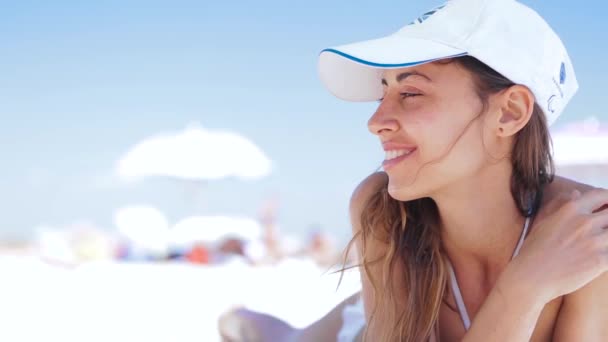 美しい日焼けした笑顔の女性が白い砂浜にビーチに横たわっている、リラックスして小さなヘッドフォンで携帯電話から音楽を聴きます。ビキニとホワイトキャップ enjoing とトプリーのスリムセクシーな女性 — ストック動画