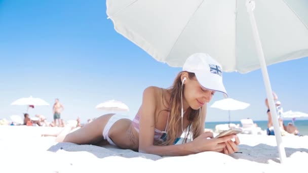 Bella abbronzata donna sorridente è sdraiata sulla spiaggia su una sabbia bianca, rilassante e ascoltare musica dal telefono con piccole cuffie. sottile donna sexy in bikini e cappuccio bianco godendo e abbronzatura su — Video Stock