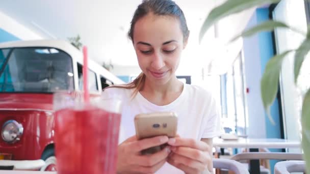 穿着白色 t恤的漂亮年轻晒黑的女人坐在咖啡馆里, 用电话、看、读或搜索东西。前景上有西瓜柠檬水的杯子 — 图库视频影像