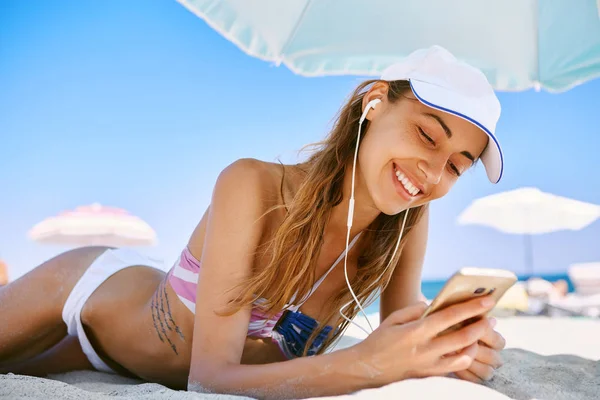 beautiful tanned smiling woman is lying on the beach on a white sand, relaxing and listen music from the phone with small headphones. slim sexy woman in bikini and white cap enjoing and suntanning on