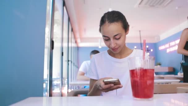 Pretty young tanned woman in white t-shirt is sitting in cafe and uses phone, looking, reading or searching something. the glass with watermelon lemonade on the foreground — Stock Video