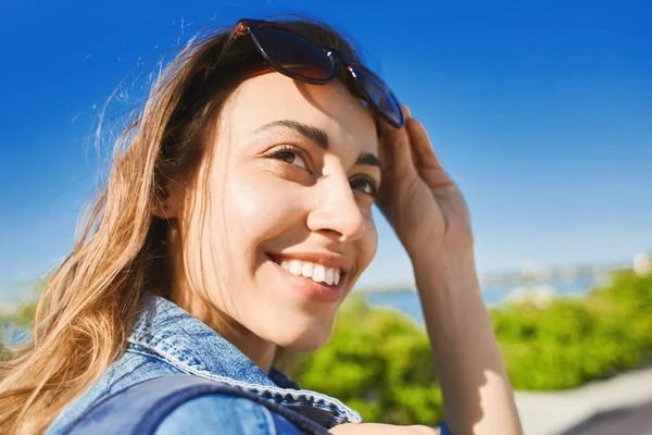 Retrato de una joven mujer atractiva sonriente con gafas de sol en un día soleado sobre el fondo azul del cielo. mujer alegre posando al aire libre —  Fotos de Stock