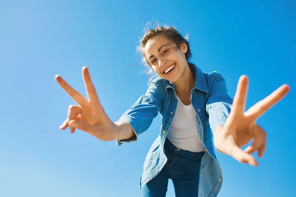 Portrait of a young smiling attractive woman in jeans clothes at sunny day on the blue sky background. woman shows a Victory sign by both hands — Stock Photo, Image