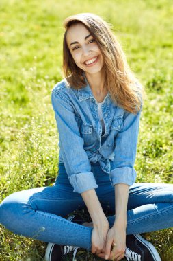 Güneşli yaz gününde çimenlerin üzerinde bir parkta otururken Jeans çekici kadın portresi.