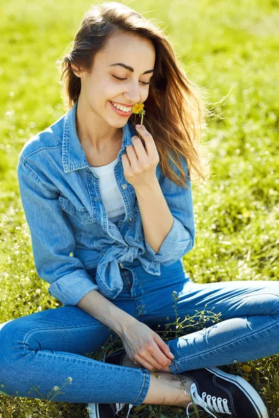 Retrato de mujer atractiva en jeans sentados en un parque en la hierba en el soleado día de verano. alegre sonriente mujer sostiene una flor de diente de león en la mano —  Fotos de Stock