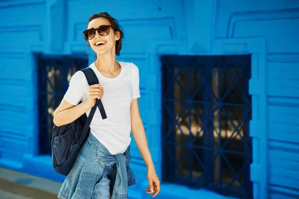 Retrato de una joven mujer atractiva sonriente en camiseta blanca con una pequeña mochila de la ciudad en un día soleado en el fondo del edificio de la ciudad. mujer posa en paisaje urbano —  Fotos de Stock