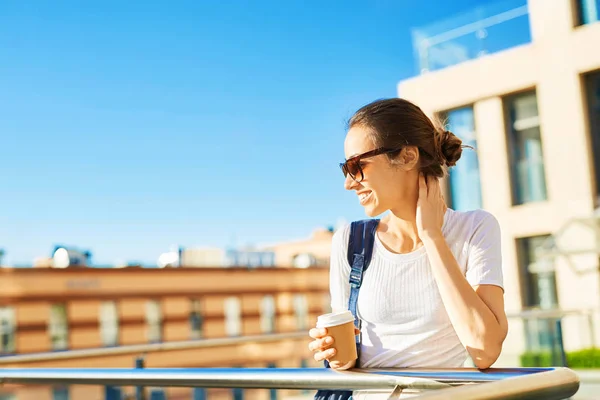 Портрет молодой улыбающейся привлекательной женщины в белой футболке с маленьким городским рюкзаком в солнечный день на фоне городского строительства. женщина позирует в городском пейзаже — стоковое фото