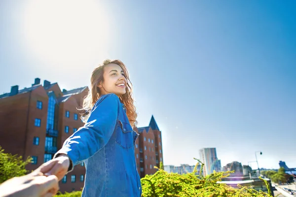 Портрет молодой улыбающейся привлекательной женщины в джинсовой одежде в солнечный день на фоне голубого неба. счастливая женщина протягивает руку кому-то вроде меня  . — стоковое фото