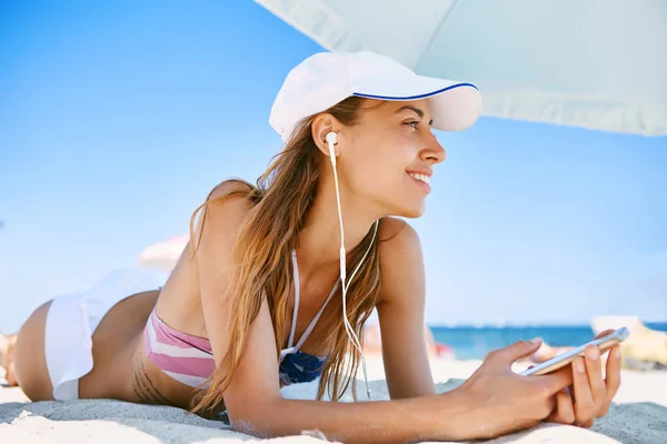 Красива засмучена усміхнена жінка лежить на пляжі на білому піску, розслабляє і слухає музику з телефону з маленькими навушниками. Тонка сексуальна жінка в бікіні і білій кепці насолоджується і засмагає на — стокове фото