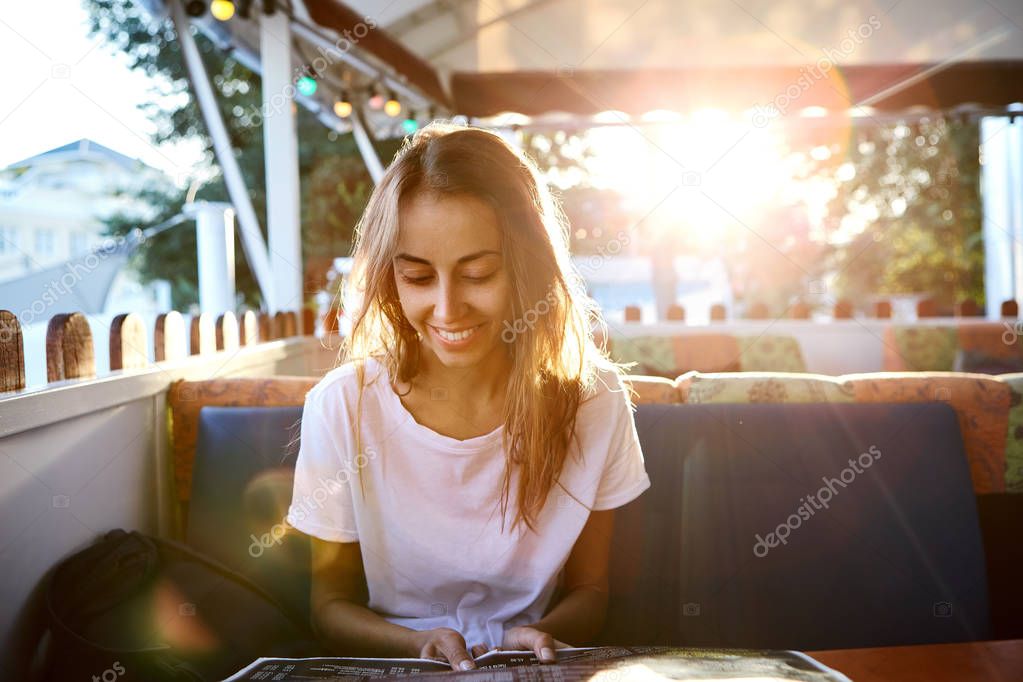 Young beautiful woman in white t-shirt sitting in outdoor cafe with sunset background.