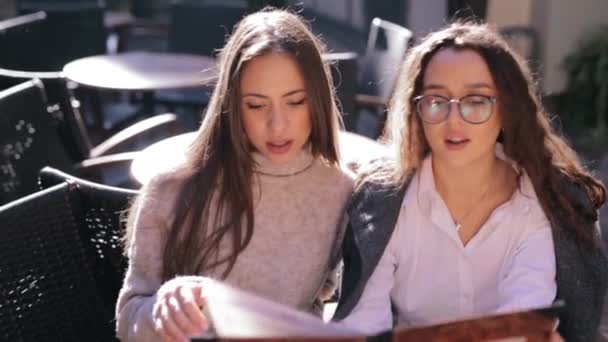 Dos mujeres hermosas jóvenes mirando el menú en el restaurante, pasando páginas y la elección de platos para el desayuno — Vídeo de stock
